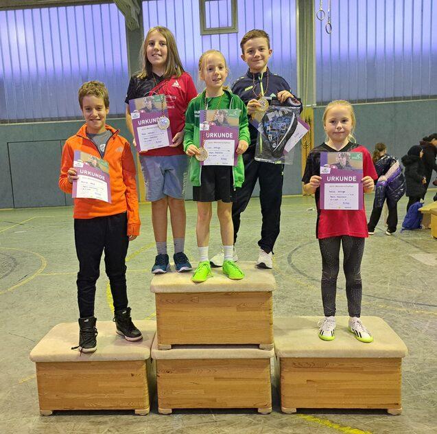 F&uuml;nf Kinder stehen auf Siegertreppchen und halten Urkunden nach einem Wettbewerb in einer Sporthalle.