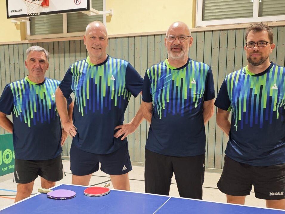 Vier Tischtennisspieler in sportlichen Shirts stehen vor einem Tischtennistisch mit roten und blauen B&auml;llen.