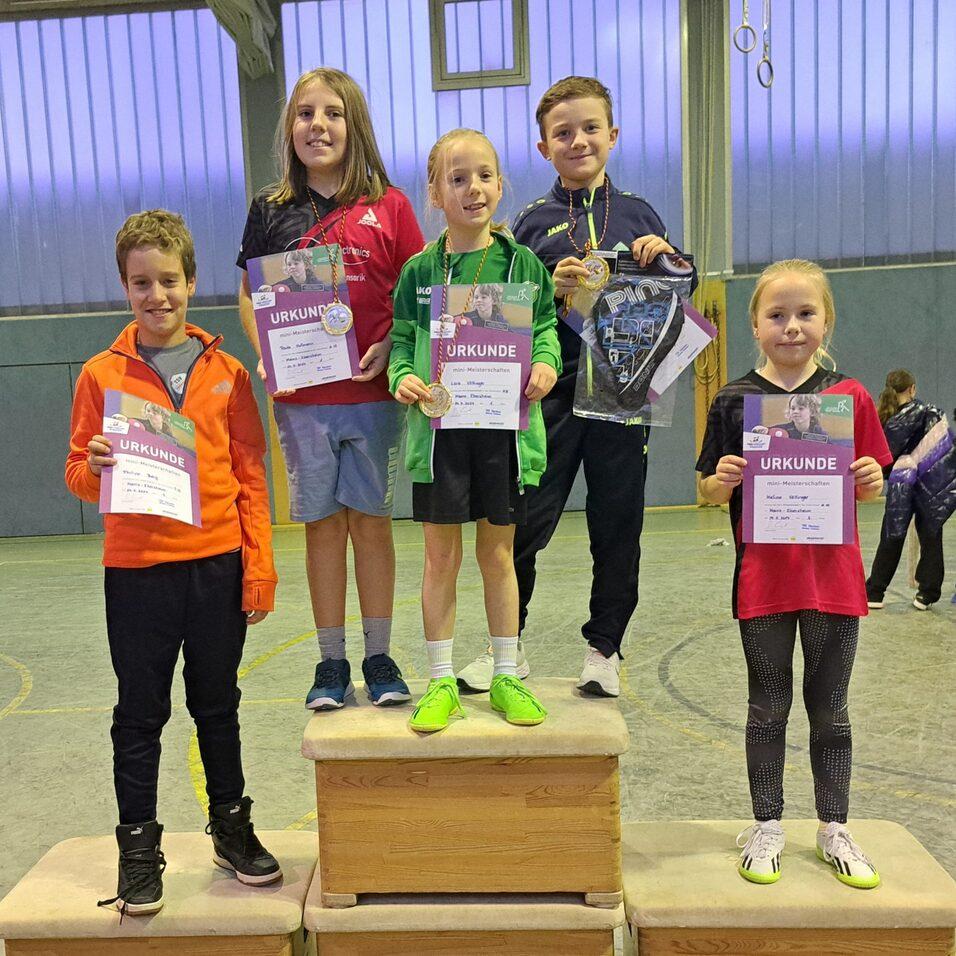 Fünf Kinder stehen auf Siegertreppchen und halten Urkunden nach einem Wettbewerb in einer Sporthalle.