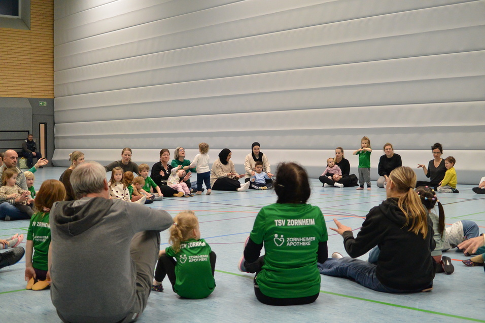 Eine Gruppe von Kindern und Erwachsenen sitzt auf dem Boden einer Sporthalle, umgeben von grauen W&auml;nden.