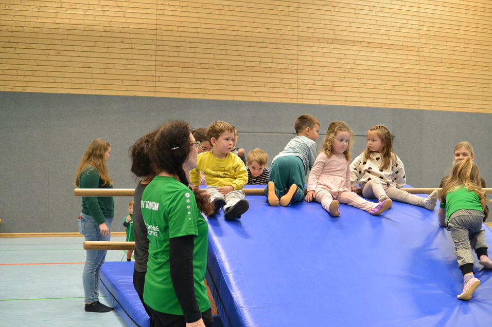 Kinder spielen auf einer blauen Gymnastikmatte in einer Sporthalle, einige klettern, andere sitzen und schauen zu.