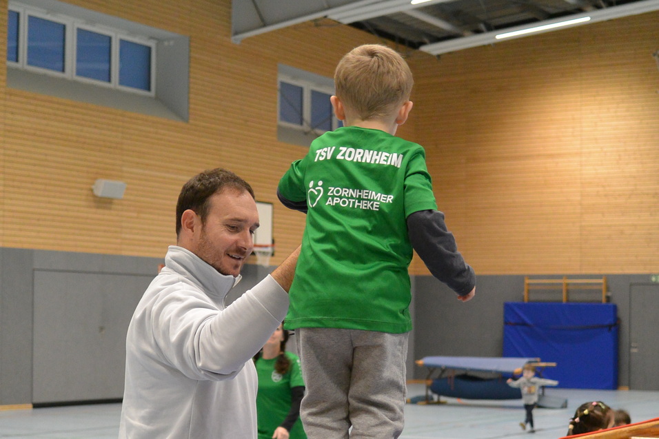 Ein Erwachsener unterst&uuml;tzt ein Kind in einem gr&uuml;nen Shirt, das auf einem Turnbock balanciert, in einer Sporthalle.