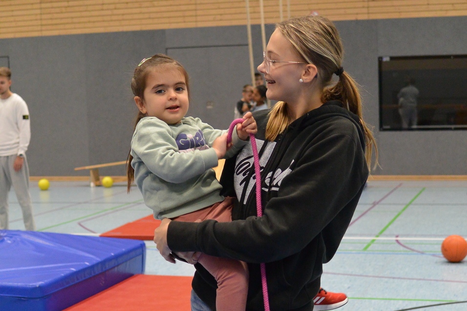 Ein Kind wird von einer jungen Frau in Sportkleidung auf dem Arm gehalten, umgeben von Turnmatten und B&auml;llen.