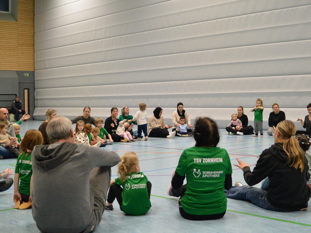 Eine Gruppe von Kindern und Erwachsenen sitzt auf dem Boden einer Sporthalle, umgeben von grauen W&auml;nden.