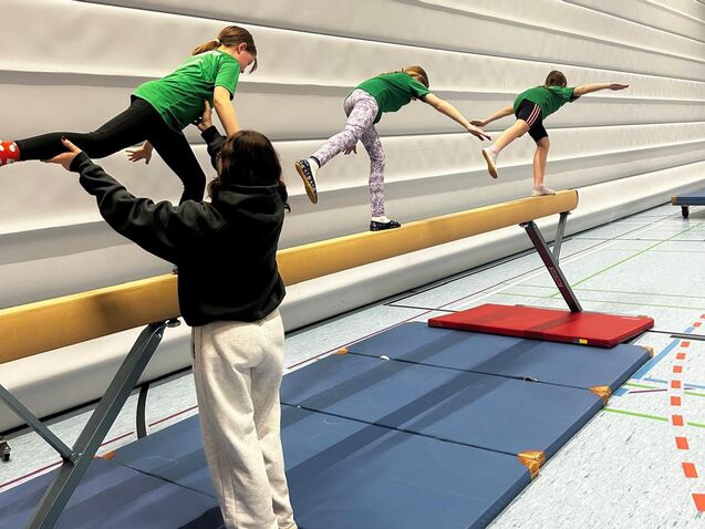 Drei Kinder balancieren auf einem Schwebebalken, w&auml;hrend ein weiteres Kind sie an der Seite unterst&uuml;tzt.