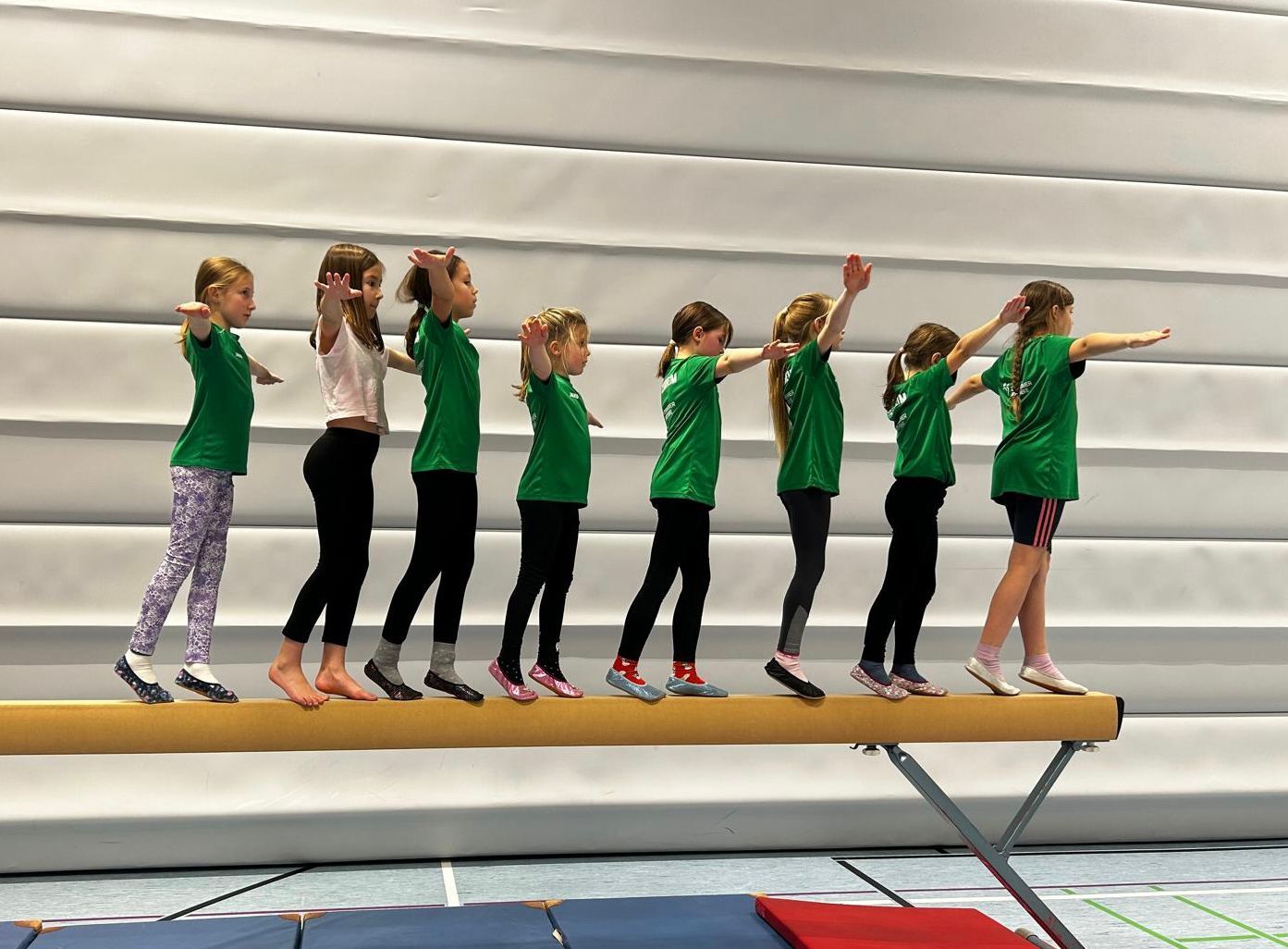 Gruppe von M&auml;dchen in gr&uuml;nen T-Shirts balanciert auf einem Schwebebalken in einer Turnhalle.