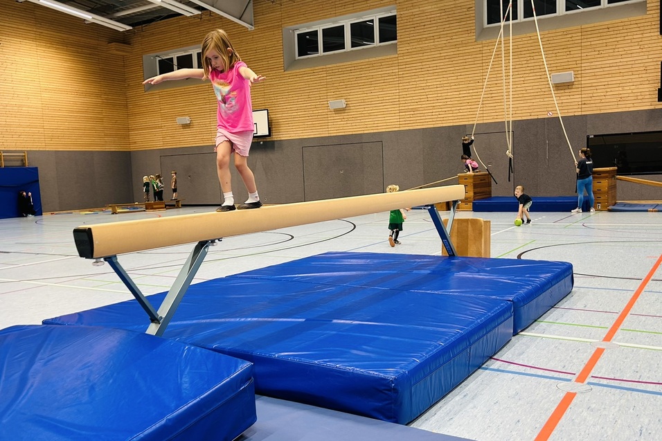 M&auml;dchen balanciert auf einem Schwebebalken in einer Sporthalle, umgeben von turnenden Kindern und Matten.