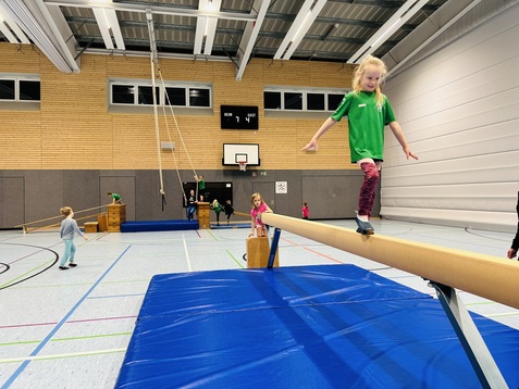 Kinder turnen in einer Sporthalle, ein M&auml;dchen balanciert auf einem Balken. Matten und K&auml;sten sind im Hintergrund sichtbar.