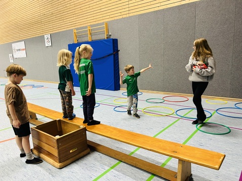 Vier Kinder in einer Sporthalle &uuml;ben auf einer Bank, w&auml;hrend eine Betreuerin mit einem Pl&uuml;schtier spricht. Reifen liegen am Boden.