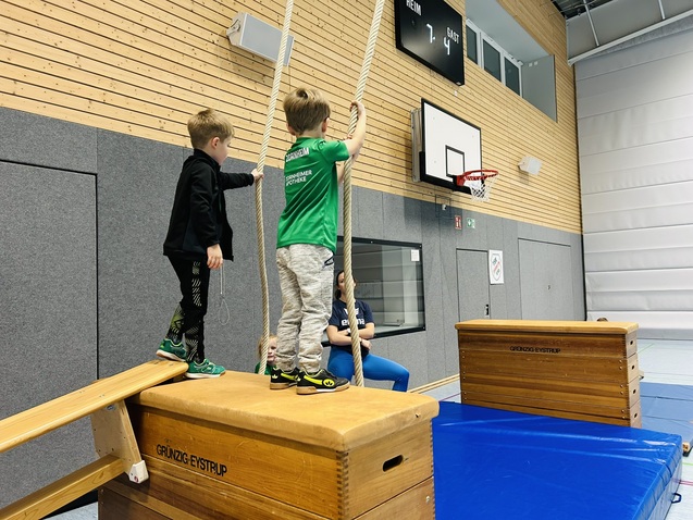 Zwei Kinder stehen auf einer Holzbank und halten sich an einem Seil in einer Sporthalle mit Turnger&auml;ten.