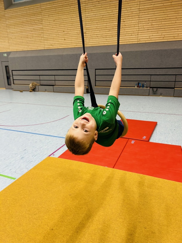 Junge in gr&uuml;nem Shirt schwingt kopf&uuml;ber an einem Schwebebalken, umgeben von bunten Turnmatten.
