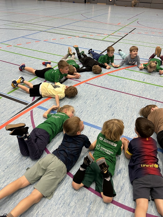 Gruppe von Kindern, die auf dem Boden einer Sporthalle liegen, in verschiedenen gr&uuml;nen T-Shirts und Shorts.