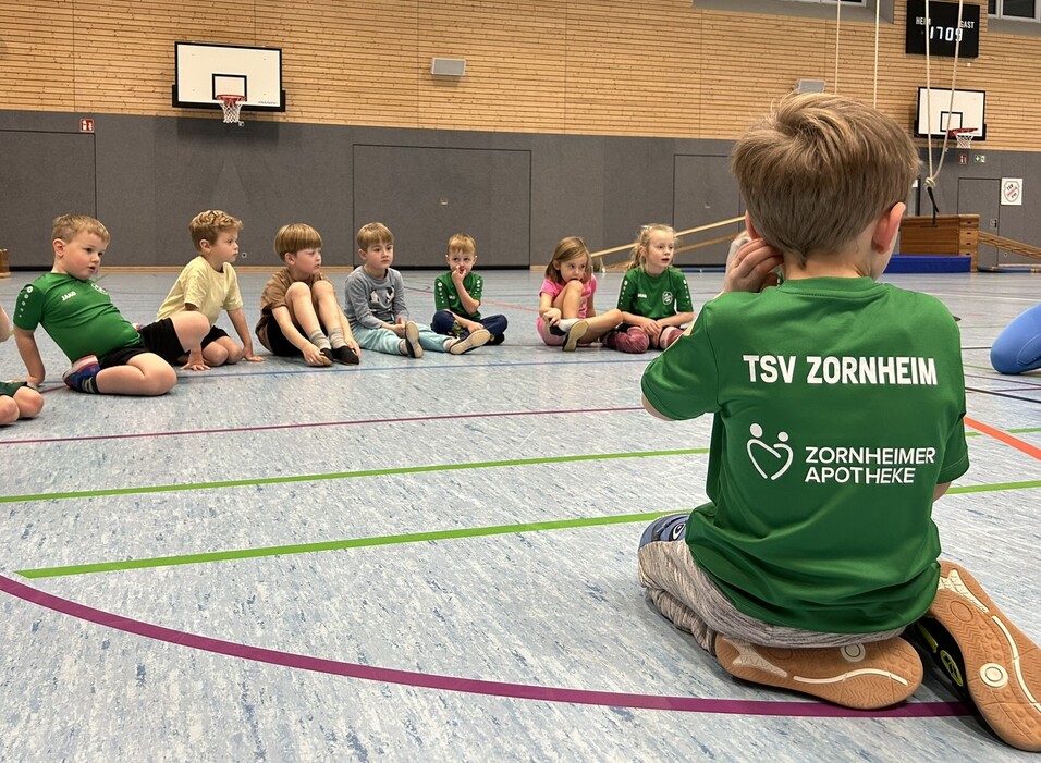 Gruppe von Kindern in Sportkleidung, die im Kreis auf dem Boden sitzen, w&auml;hrend ein Junge vorne spricht.