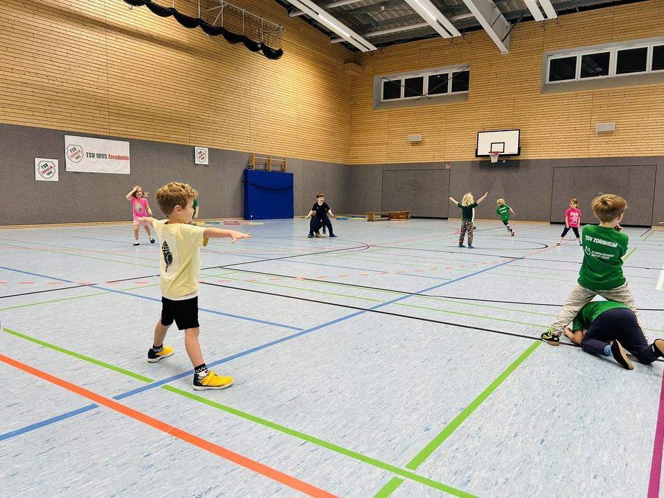 Kinder spielen in einer Sporthalle, einige laufen, andere machen &Uuml;bungen, bunt gekleidet auf einem blauen Boden.