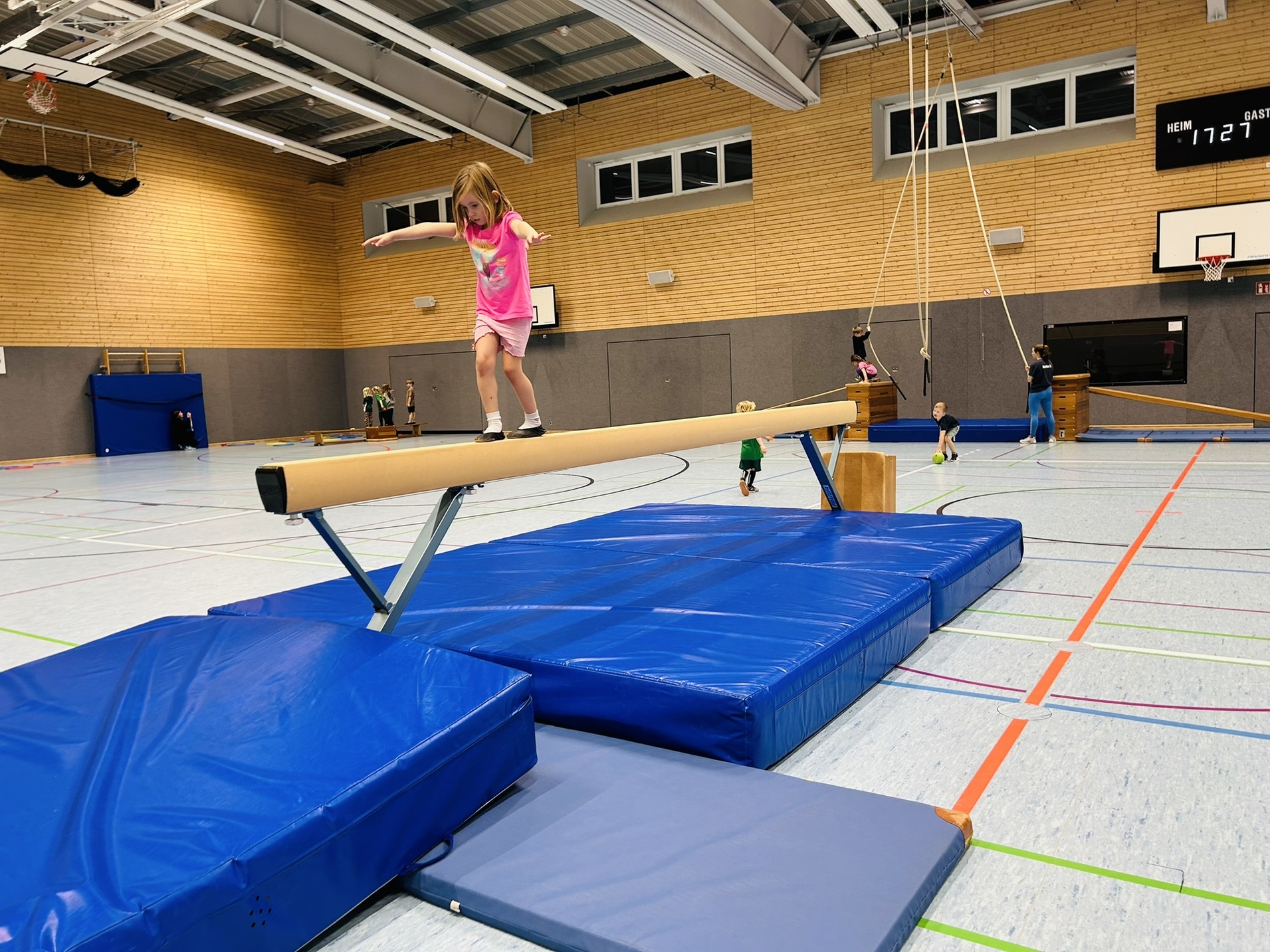 M&auml;dchen balanciert auf einem Schwebebalken in einer Sporthalle, umgeben von turnenden Kindern und Matten.