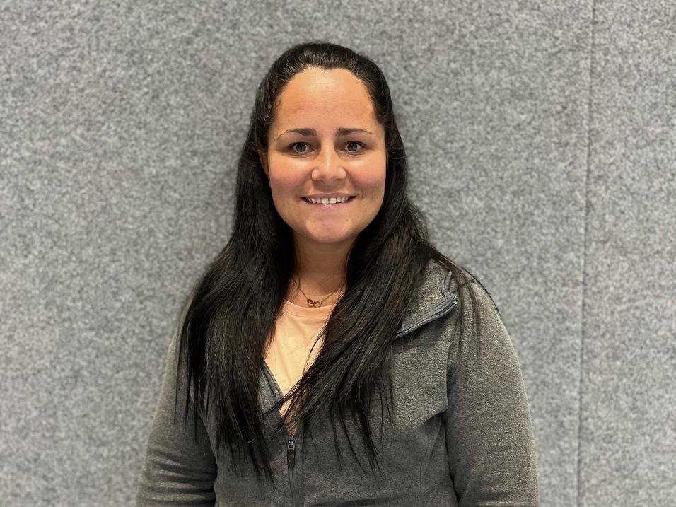 Lächelnde Frau mit langen, dunklen Haaren in grauer Jacke vor einer grauen Wand.