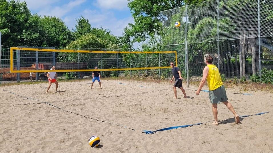 Vier Personen spielen Beachvolleyball auf einem Sandplatz, w&auml;hrend ein Kind mit einer Kappe im Hintergrund sitzt.