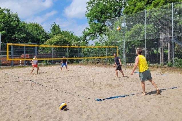 Vier Personen spielen Beachvolleyball auf einem Sandplatz, w&auml;hrend ein Kind mit einer Kappe im Hintergrund sitzt.