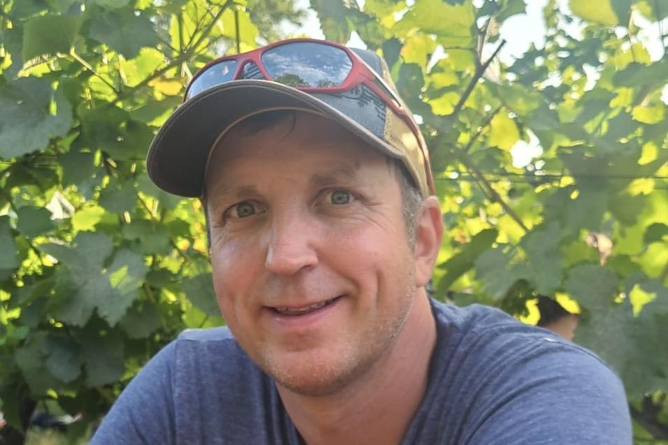 Mann mit kurzen Haaren und einer Schirmm&uuml;tze l&auml;chelt vor einem Weinberg mit gr&uuml;nen Bl&auml;ttern.