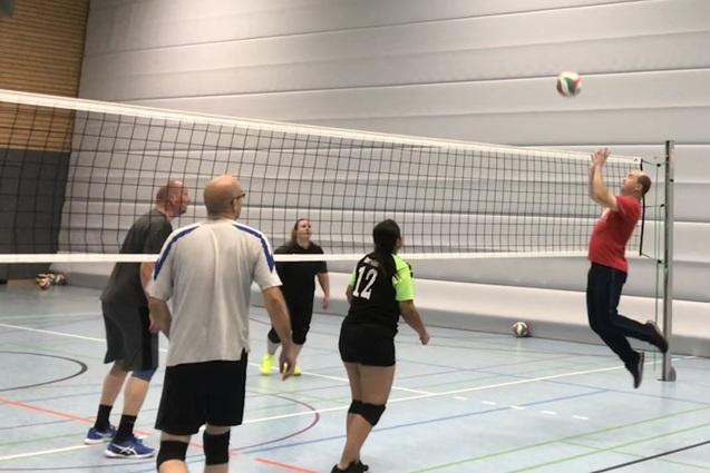 Volleyballspiel in einer Halle, ein Spieler springt zum Angriff, w&auml;hrend andere warten. Netz und Ball sichtbar.