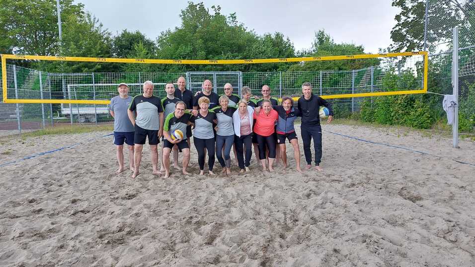 Gruppe von 15 Personen steht im Sand am Beachvolleyballfeld, hinter ihnen das Netz und B&auml;ume im Hintergrund.