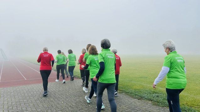 Gruppe von Frauen in gr&uuml;nen und roten Shirts beim langsamen Gehen auf einem nebeligen Sportplatz.