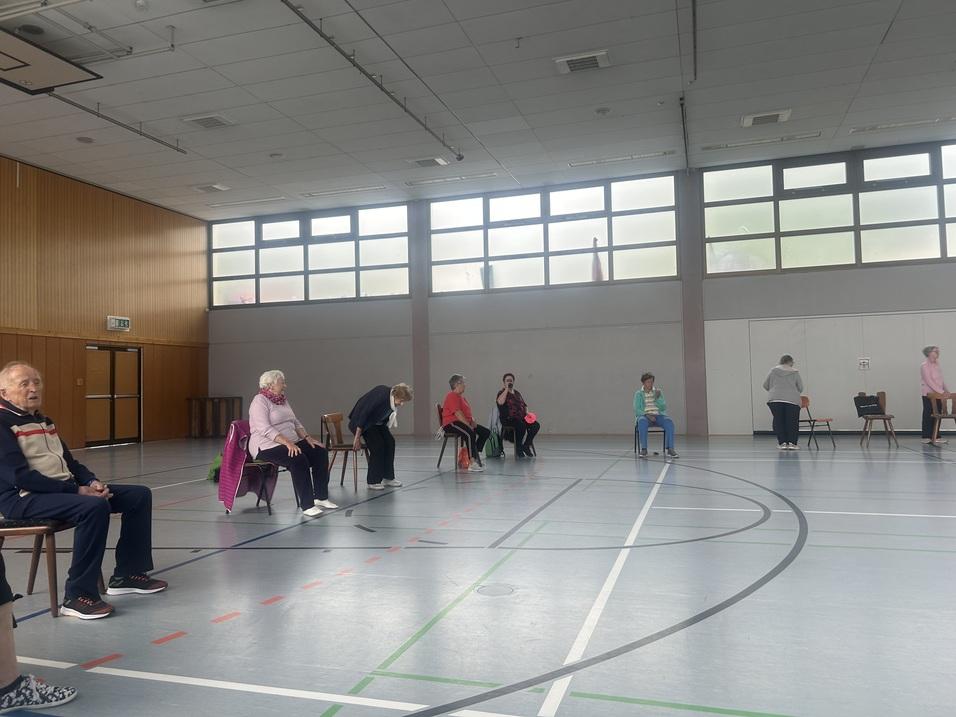 Gruppensitzung älterer Menschen in einem Sportraum, die auf Stühlen sitzen und verschiedenen Aktivitäten nachgehen.