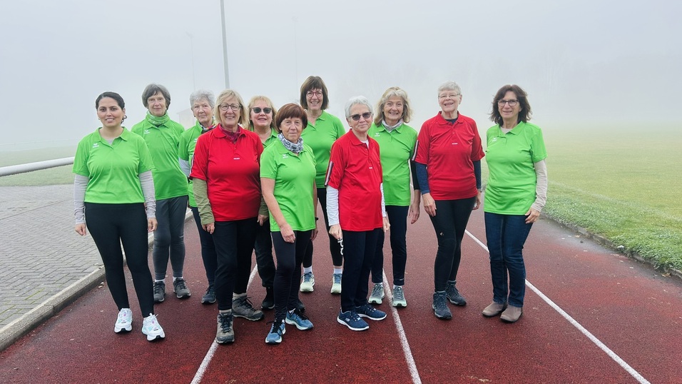 Gruppe von zehn Frauen in gr&uuml;nen und roten T-Shirts steht auf einer Laufbahn im Nebel.