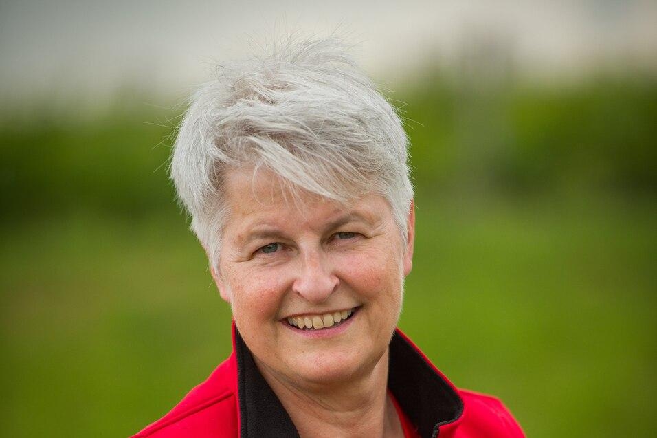 Lächelnde Frau mit kurzem, grauem Haar in rotem Poloshirt und Weste, vor einer grünen Landschaft.