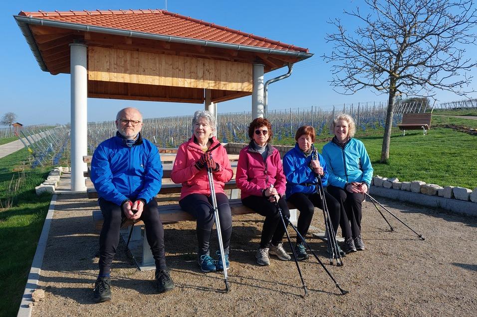 Gruppe von f&uuml;nf Personen mit Nordic-Walking-St&ouml;cken sitzt unter einem Pavillon auf einem Wegen in einer Weinlandschaft.