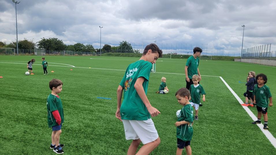 Gruppe von Kindern in gr&uuml;nen Trikots auf einem Fu&szlig;ballplatz, einige &uuml;ben mit B&auml;llen, andere stehen daneben.