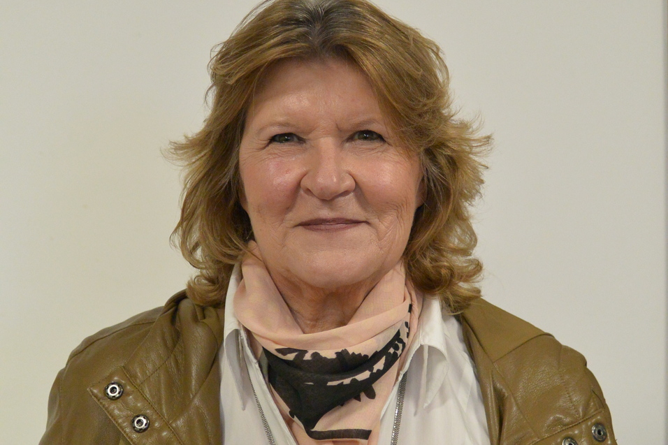 Portr&auml;t einer Frau mit blonden, lockigen Haaren, die einen braunen Mantel und einen schwarzen Schal tr&auml;gt.