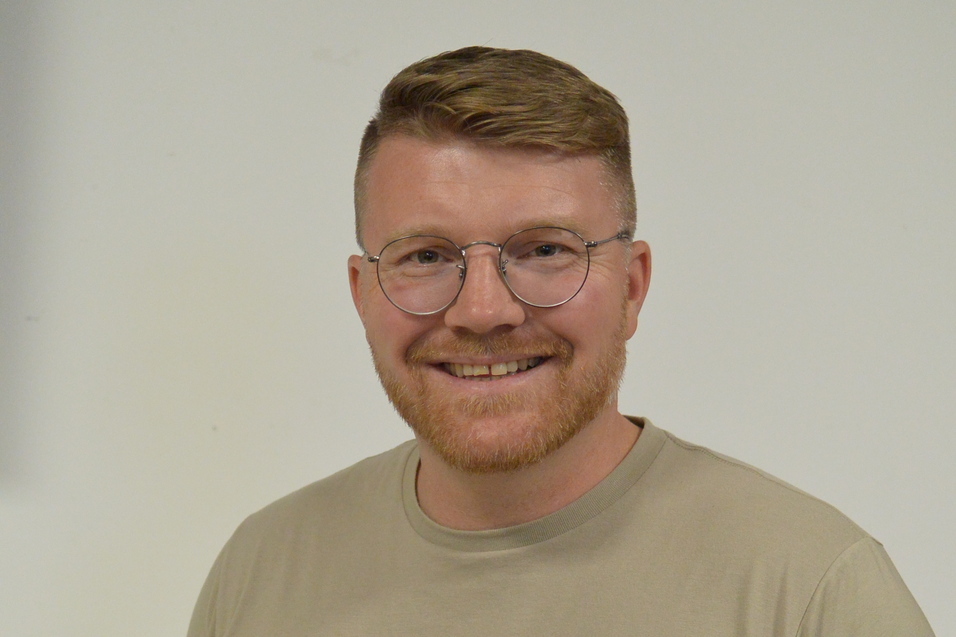 L&auml;chelnder Mann mit Brille tr&auml;gt ein beige T-Shirt vor einem hellen Hintergrund.