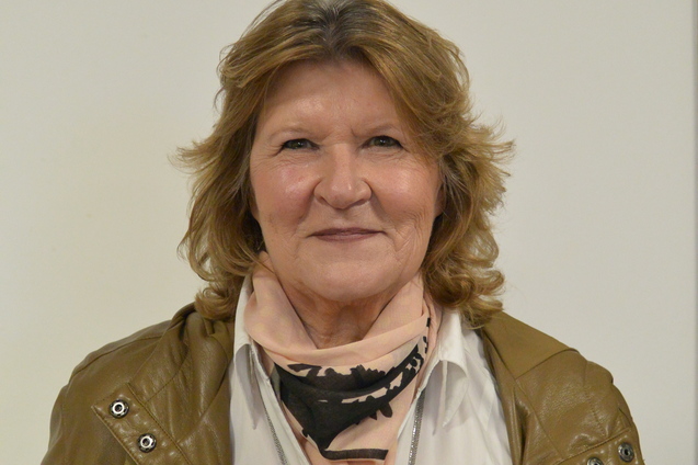 Portr&auml;t einer Frau mit blonden, lockigen Haaren, die einen braunen Mantel und einen schwarzen Schal tr&auml;gt.