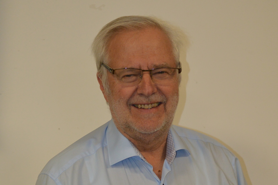 Portrait eines l&auml;chelnden &auml;lteren Mannes mit Brille, in einem hellblauen Hemd vor einem einfarbigen Hintergrund.