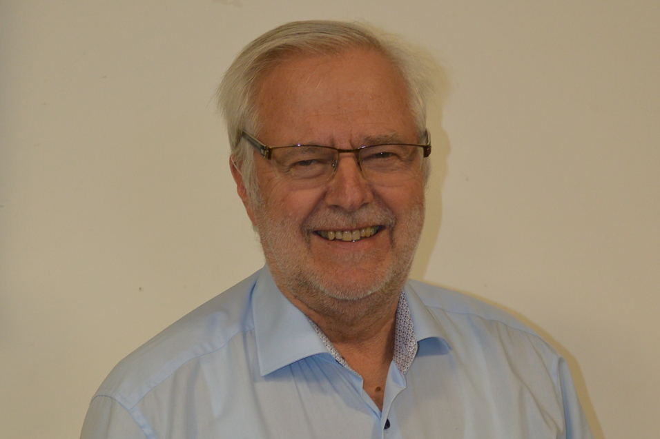 Portrait eines l&auml;chelnden &auml;lteren Mannes mit Brille, in einem hellblauen Hemd vor einem einfarbigen Hintergrund.