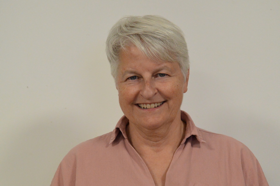 L&auml;chelnde Frau mit kurzen, grauen Haaren in einem rosafarbenen Hemd vor einem hellen Hintergrund.