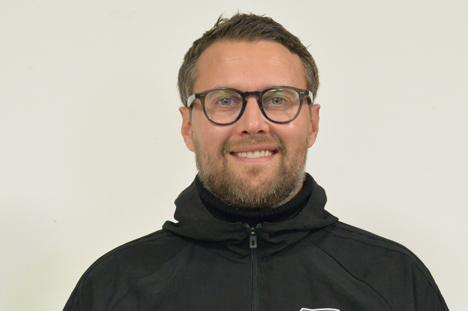 Mann mit Brille und Bart tr&auml;gt eine schwarze Jacke mit Vereinslogo vor einem neutralen Hintergrund.