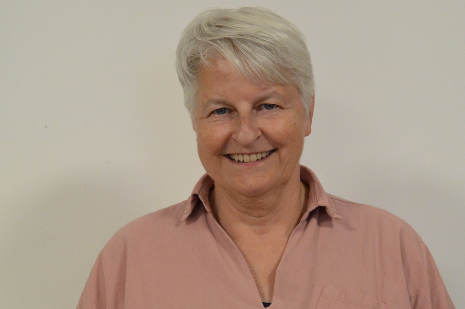 L&auml;chelnde Frau mit kurzen, grauen Haaren in einem rosafarbenen Hemd vor einem hellen Hintergrund.