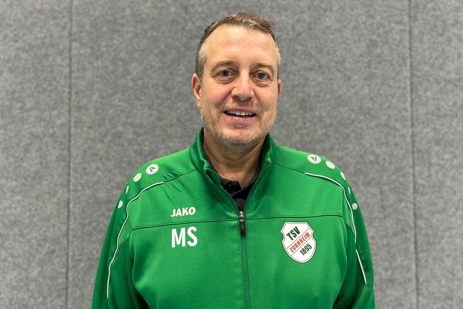 Ein Mann in einem gr&uuml;nen Sportjacke mit dem Schriftzug "TSV" und Initialen "MS" steht vor einem grauen Hintergrund.