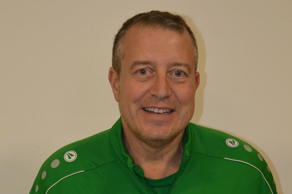 Portr&auml;t eines l&auml;chelnden Mannes in einem gr&uuml;nen Trainingsanzug mit Logo und Initialen, vor einer hellen Wand.