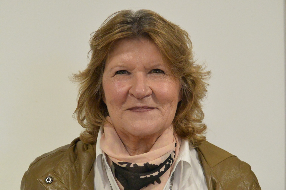 Portr&auml;t einer Frau mit blonden, lockigen Haaren, die einen braunen Mantel und einen schwarzen Schal tr&auml;gt.