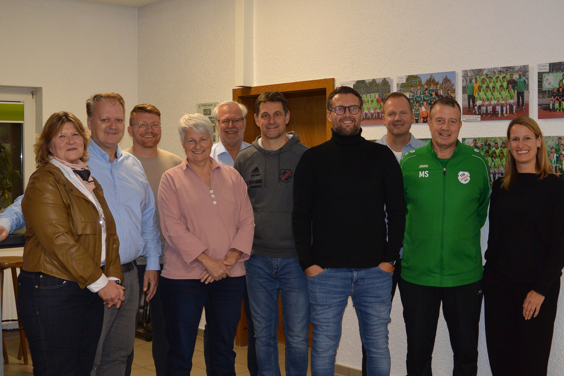 Gruppendfoto von elf Personen in sportlicher Umgebung, l&auml;chelnd, vor W&auml;nden mit Teamfotos.