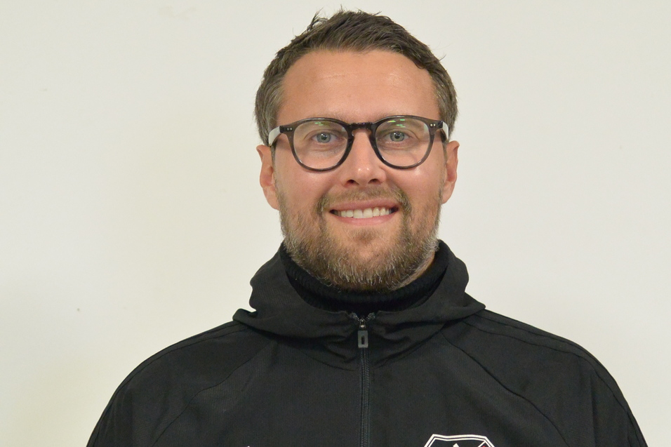 Mann mit Brille und Bart tr&auml;gt eine schwarze Jacke mit Vereinslogo vor einem neutralen Hintergrund.