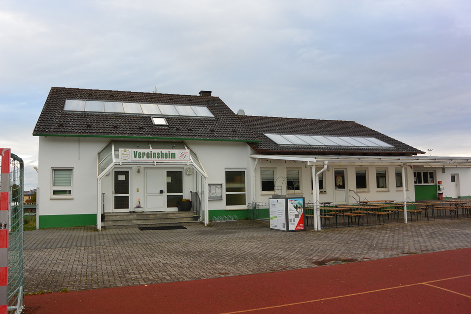 Vereinsheim mit Terrasse und Solarpanelen, umgeben von einem geschotterten Platz und Sportanlagen.