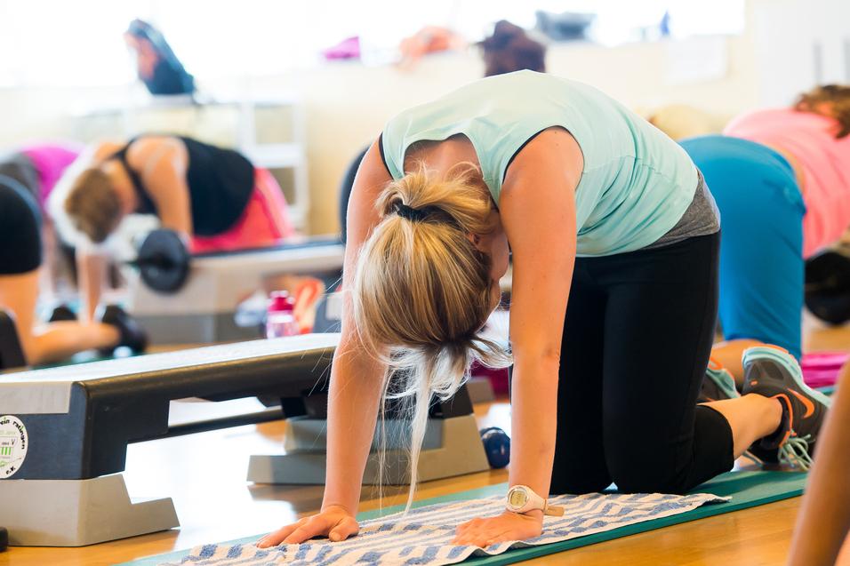 Frau in Fitnesskleidung übt auf ihrer Matte in einem Gruppenfitnesskurs mit weiteren Teilnehmern im Hintergrund.