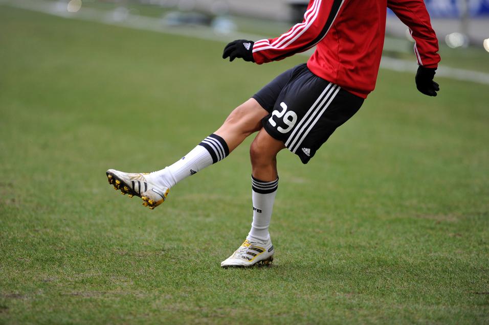 Nahaufnahme eines Fußballspielers mit einem roten Trikot, der auf einem Rasenfeld einen Ball mit dem rechten Fuß spielt.