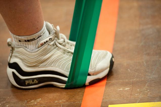 Nahaufnahme eines Fußes in einem grauen Sportschuh, der von einem grünen Widerstandsband gehalten wird.